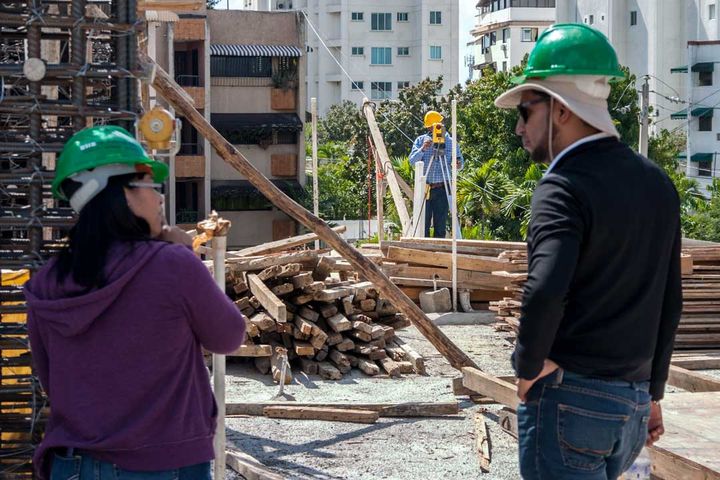 Two people from the firm watch a third take a reading with the level, among stacks of formwork lumber