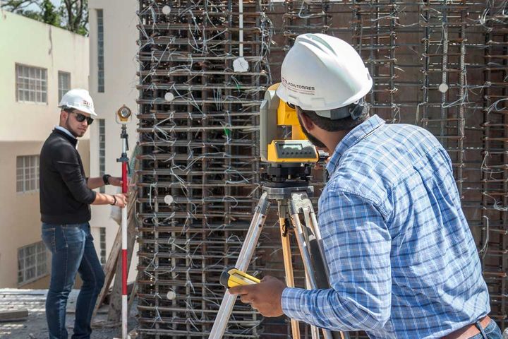 Optical level on a tripod in front of a column rebar cage, with two members of the firm