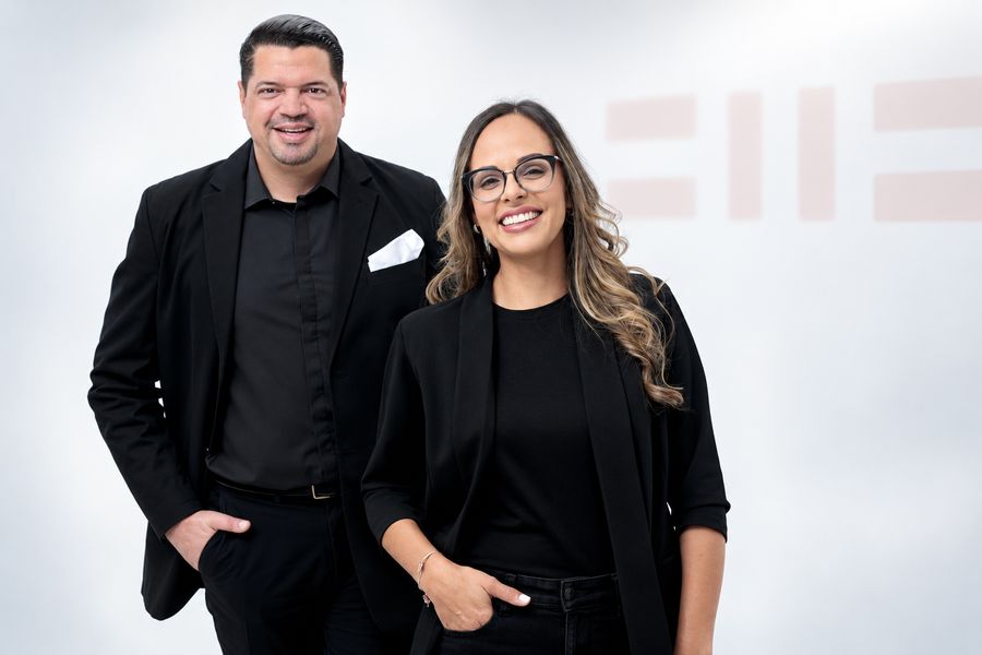 Andrés Torres and Mariel Tejada standing and smiling, dressed in black, in front of a light background with the firm’s logo